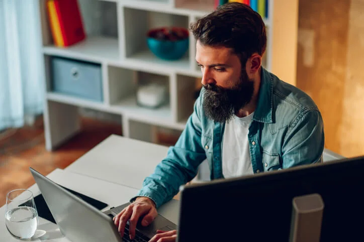 A young focused bearded writer is sitting at home and typing an article on a laptop. A writer in smart casual is sitting at home and typing a report on a laptop while working remotely.